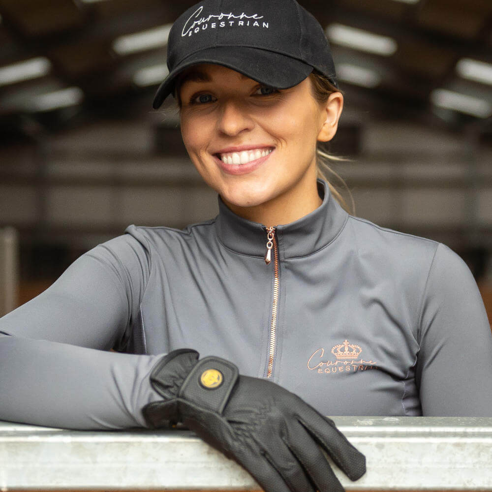 Equestrian rider with a vibrant smile and equestrian clothing from McCaskie.store, leaning on a gate at the horse stables. Find high-quality equestrian wear and gear at McCaskie.store, your one-stop-shop for all your horse riding needs.