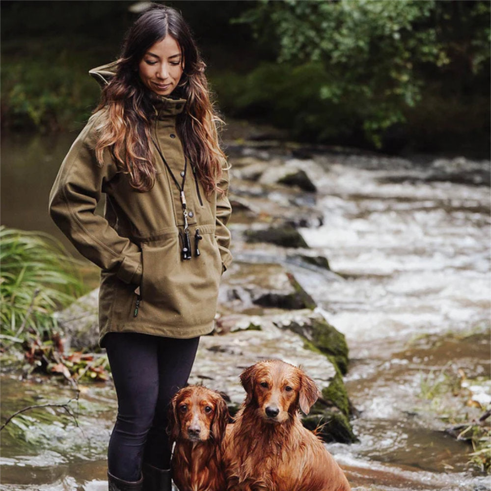 Beautiful brunette woman wearing a Ridgeline jacket with her dogs by a river in a forest. Dress for any weather and outdoor activity with high-quality and durable clothing from Ridgeline of New Zealand. Visit McCaskie.store website today get yours!